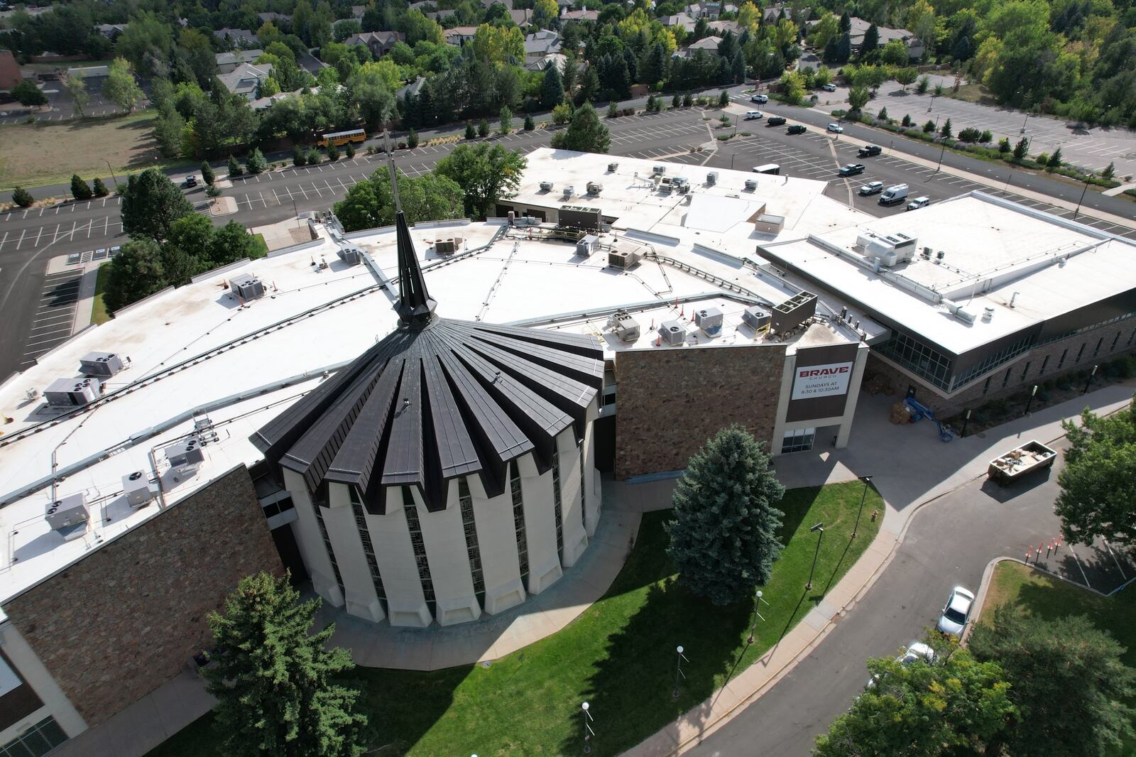 Aerial view of a church campus with white flat membrane roofs around a fluted metal sanctuary roof and spire